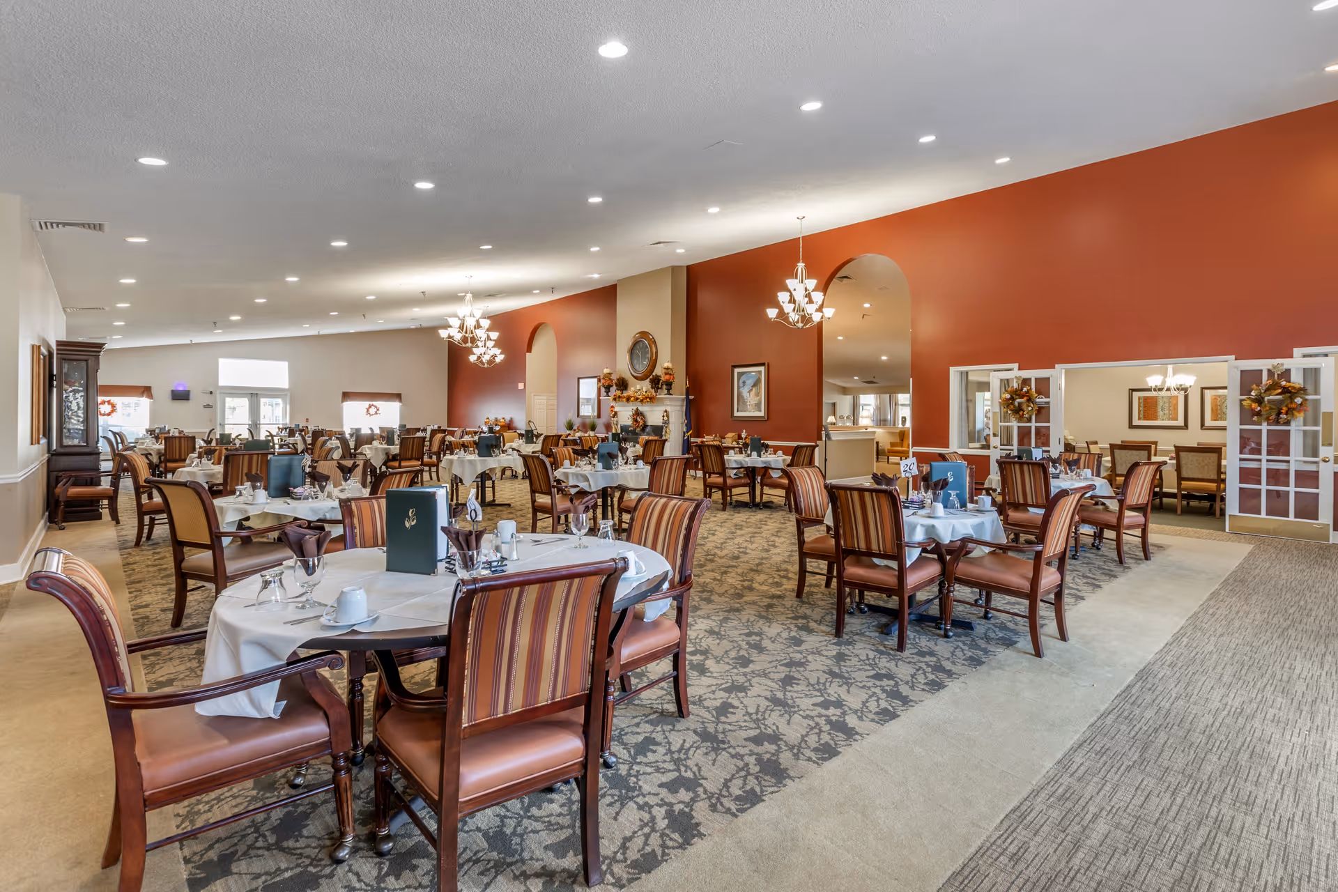 A spacious dining room in a senior living facility with multiple round tables covered with white tablecloths, set with cups, glasses, and menus. The room features striped cushioned chairs, chandeliers, a red accent wall with a fireplace decorated for fall, and large windows letting in natural light.