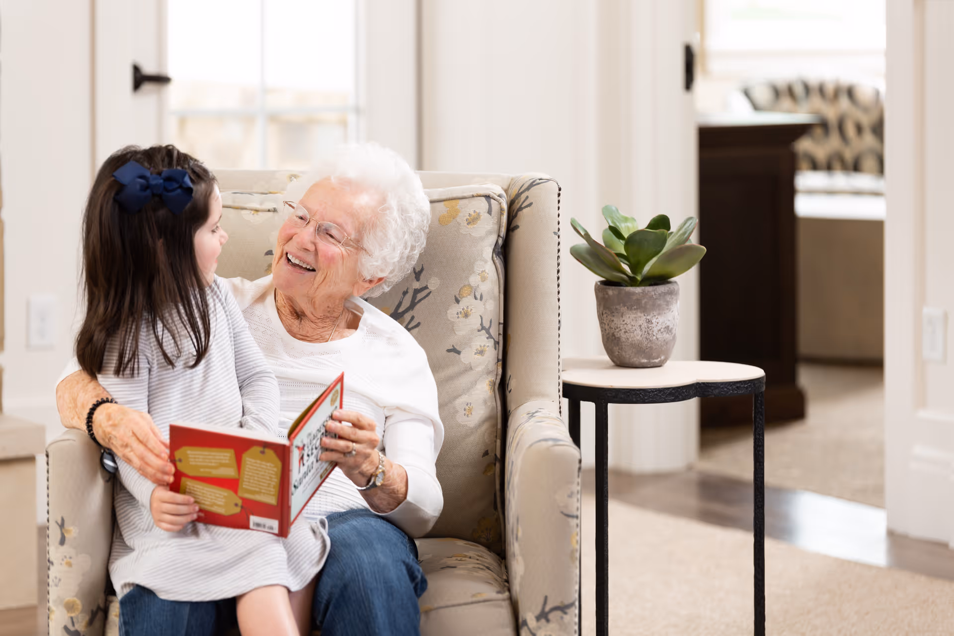 An elderly woman sits in an armchair smiling as she reads a book to a young girl on her lap in a bright living area.