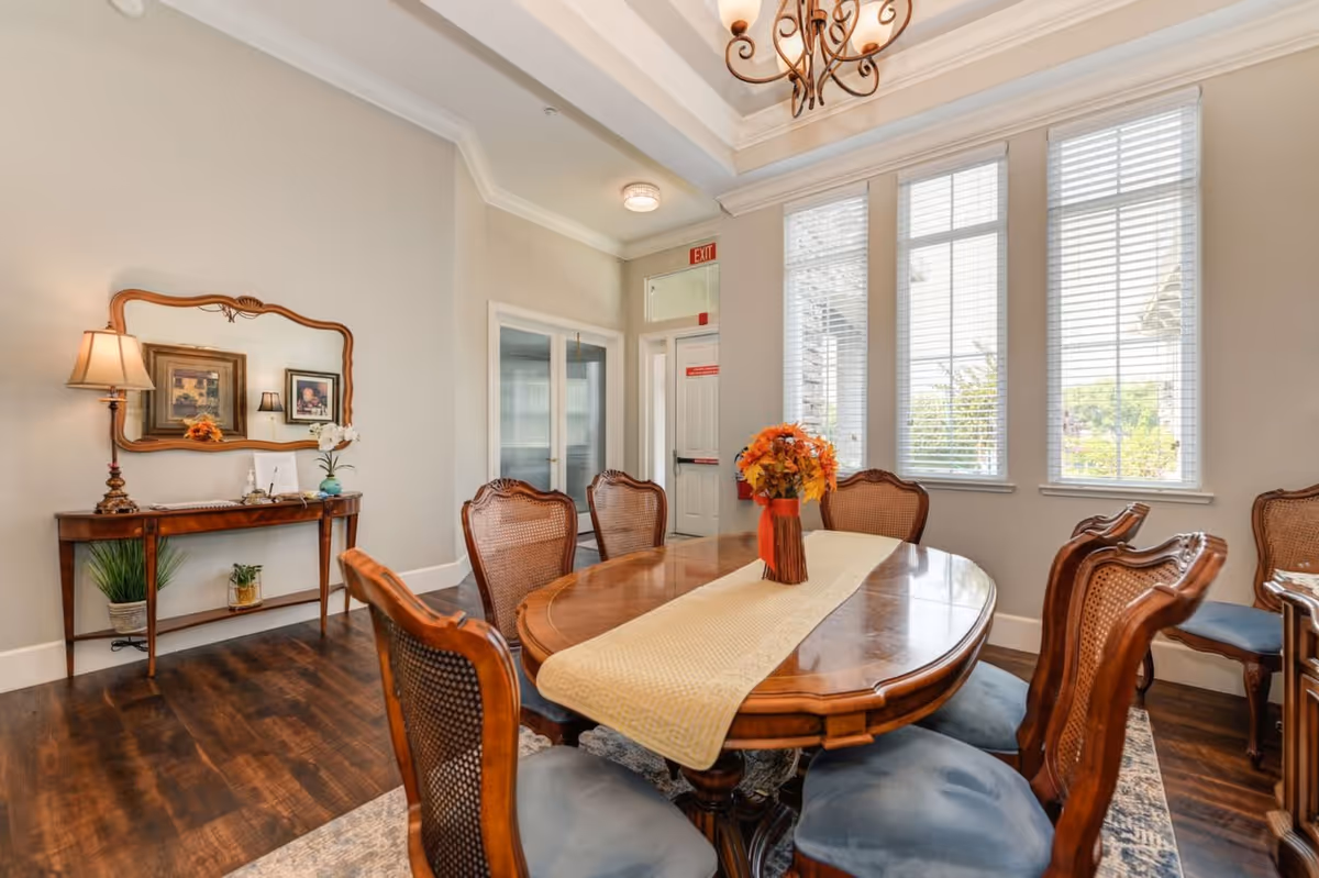 A well-lit dining room with a wooden dining table surrounded by six cushioned chairs. The table has a beige runner and a vase with orange flowers. There are three large windows with white blinds letting in natural light. On the left wall, there is a wooden console table with a lamp, a mirror, and framed pictures above it. The floor is dark wood, and the walls are painted light beige.