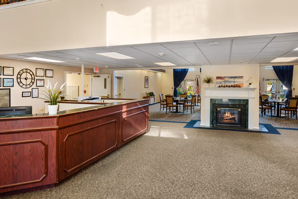 Reception area with a wooden front desk and a potted plant on it. Behind the desk, there are framed certificates and a large wall clock. In the background, there is a seating area with tables and chairs, a fireplace with a fire burning, and windows with blue curtains letting in natural light.