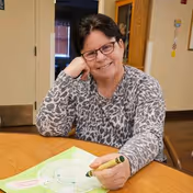 An elderly woman with glasses and short dark hair is sitting at a wooden table, smiling and holding a green crayon while coloring a drawing on a piece of paper. She is wearing a gray patterned long-sleeve shirt. The background shows an interior room with a door and some wall decorations.