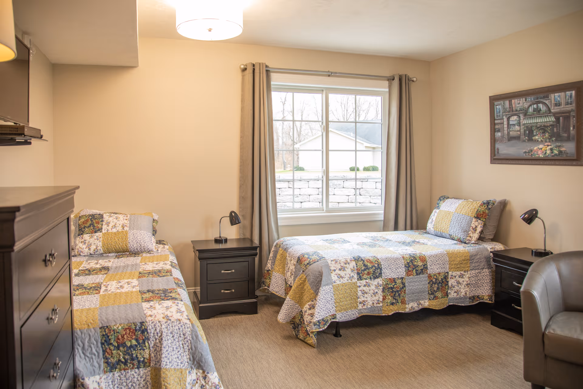A cozy bedroom with two twin beds covered in patchwork quilts in shades of yellow, blue, and floral patterns. Each bed has a matching pillow. Between the beds is a black nightstand with a small lamp. There is a large window with beige curtains letting in natural light. On the right side of the room is a framed picture on the wall and a gray armchair. A black dresser is partially visible on the left side of the image. The room has beige walls and carpeted flooring.