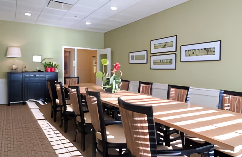A well-lit dining room with a long wooden table surrounded by chairs. The walls are painted light green and decorated with four framed pictures. There is a sideboard with a lamp and plants in the background, and sunlight streams through windows casting shadows on the table and floor.