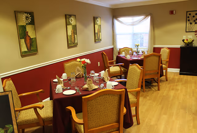 A dining room in a senior living community with two tables covered in burgundy tablecloths, each set with plates, cups, glasses, and folded napkins. There are six upholstered wooden chairs around the tables. The room has light wood flooring, beige walls with a red lower half, and three framed floral paintings on the wall. A window with sheer curtains lets in natural light, and a dark wooden cabinet with a bowl of fruit is in the corner.