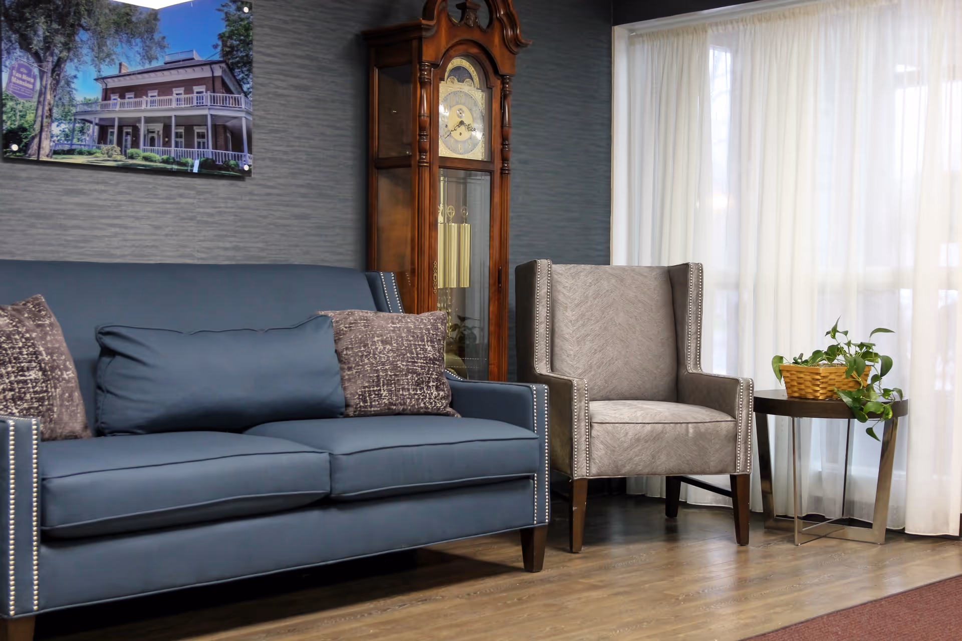 A cozy sitting area with a blue sofa adorned with three cushions, a gray upholstered armchair, a wooden grandfather clock, a small side table with a basket of green plants, and sheer white curtains covering a large window.