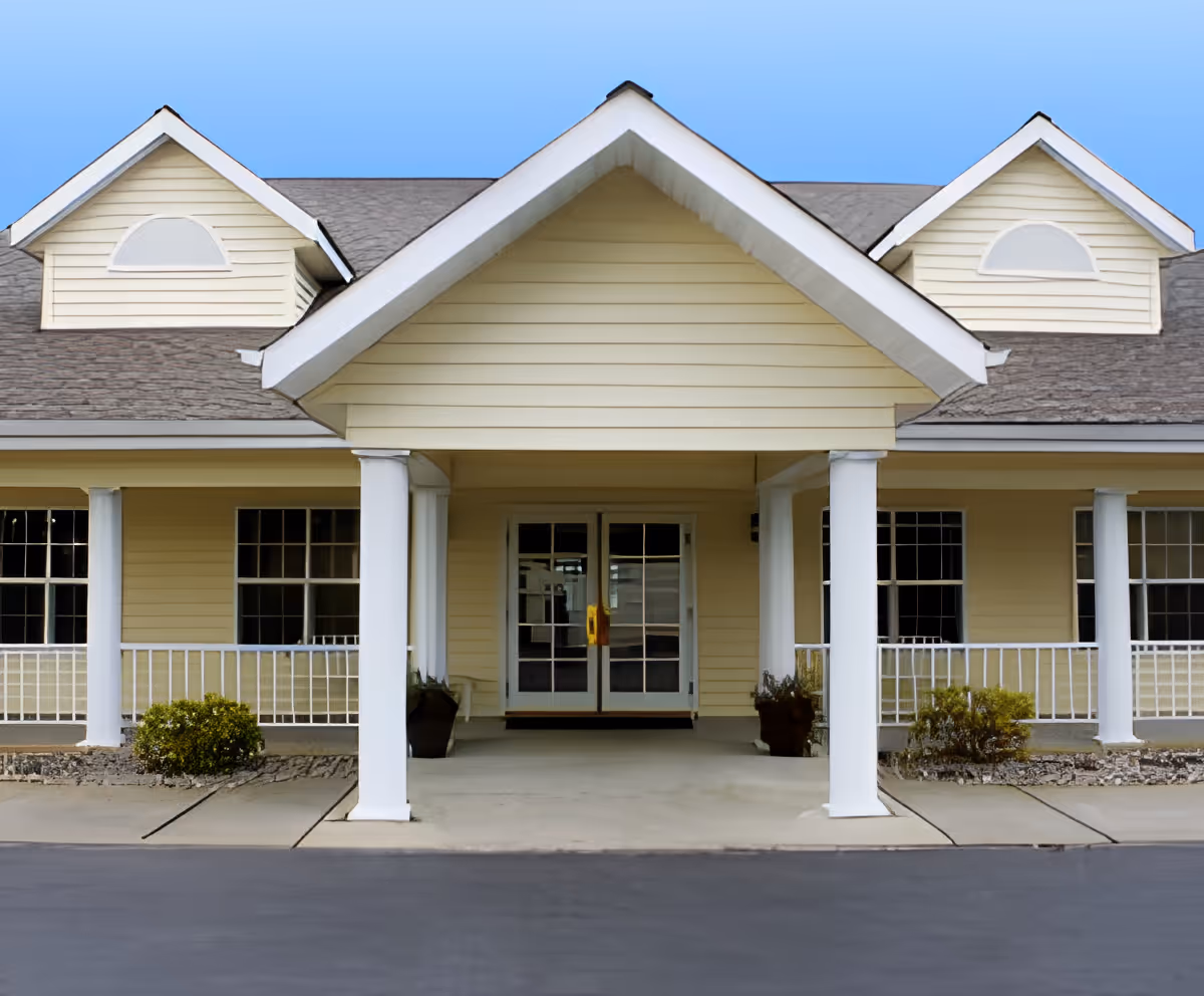 Front exterior view of a single-story building with a covered entrance supported by white columns, beige siding, and two dormer windows on the roof under a clear blue sky.