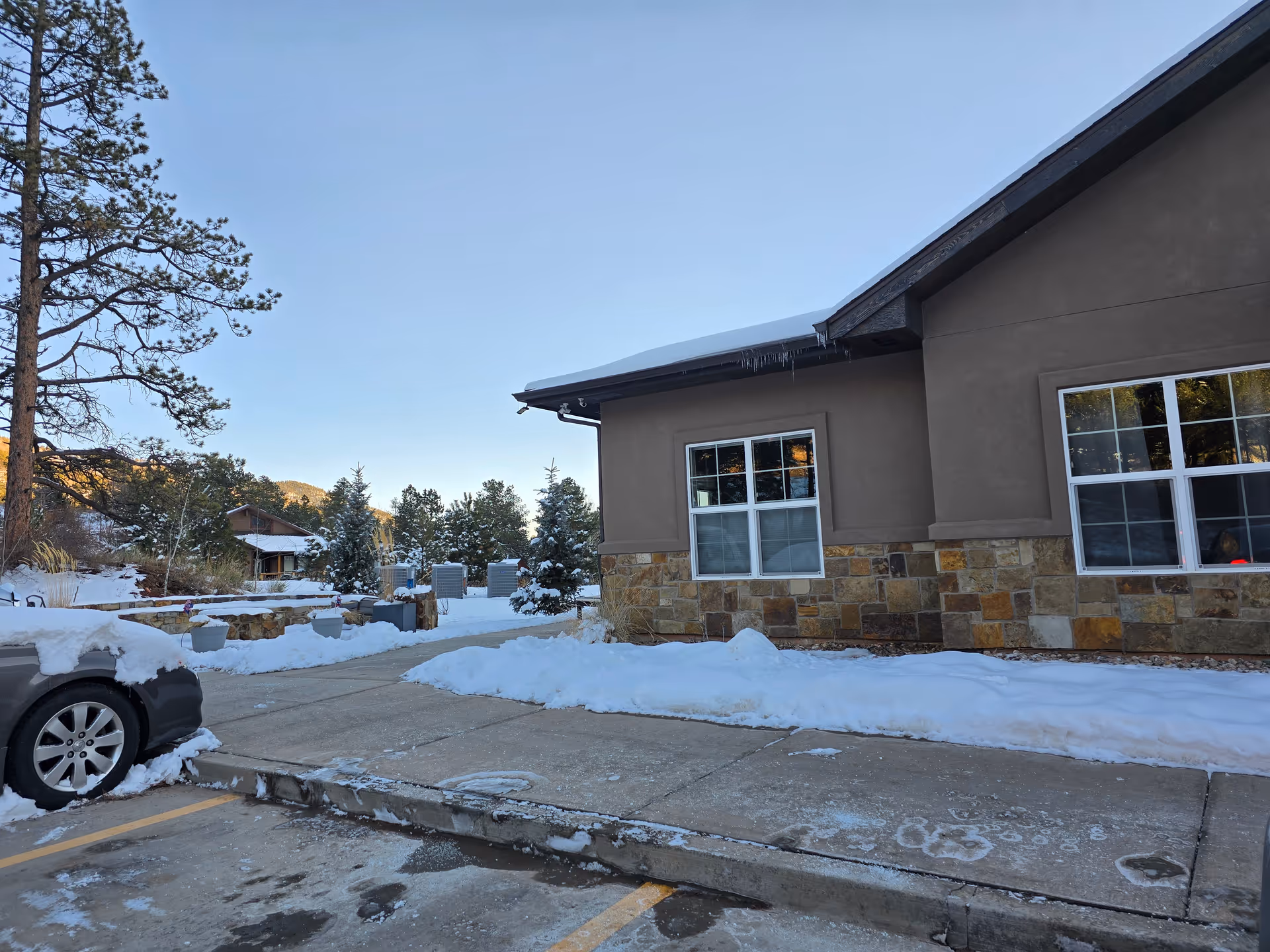 Snow-covered parking area and sidewalk in front of a stone-accented building with windows and trees in the background.
