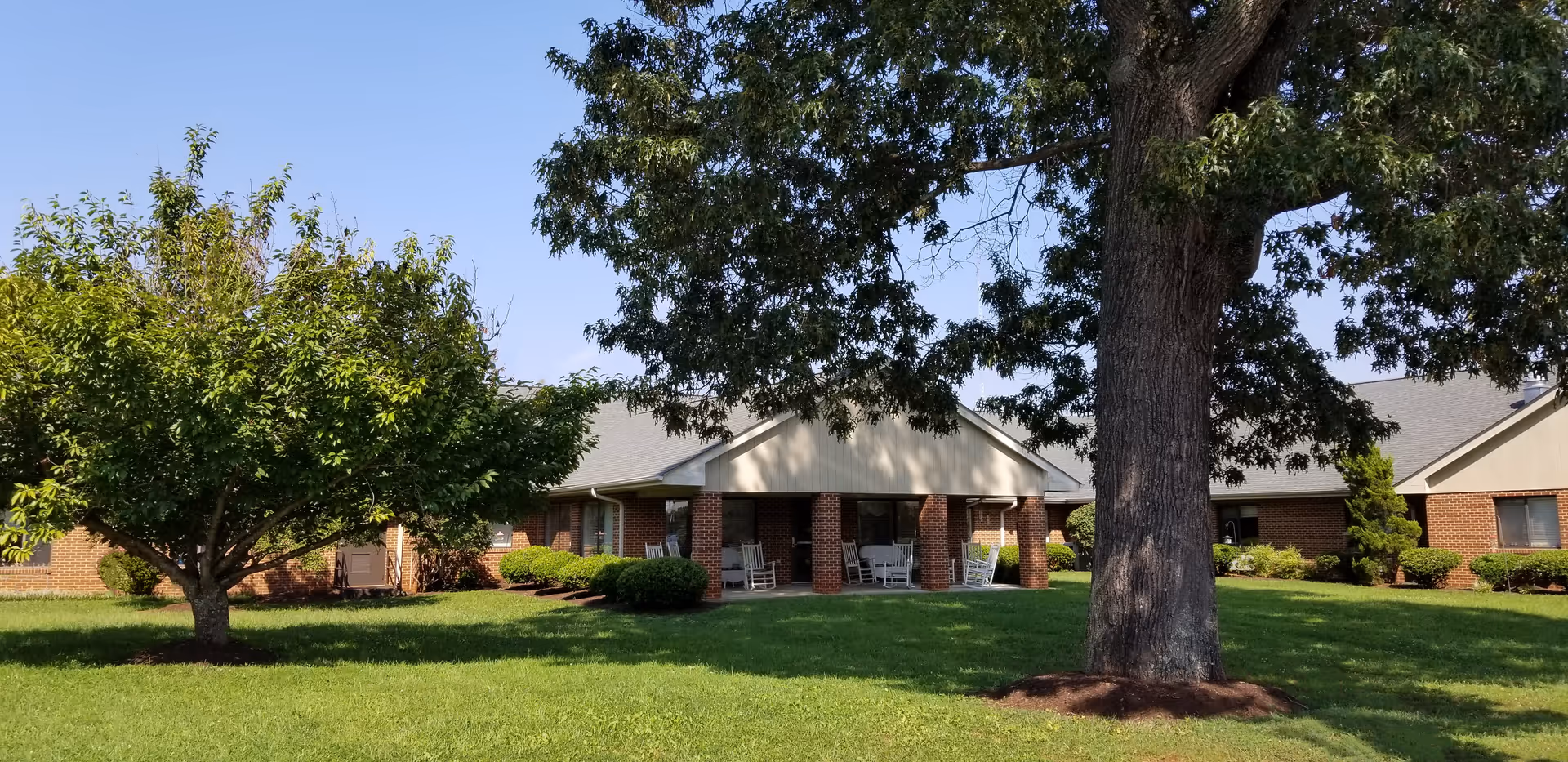 A single-story brick building with a covered porch area featuring several white rocking chairs. The building is surrounded by a well-maintained grassy lawn with a large tree in the foreground and another smaller tree to the left. The sky is clear and blue.