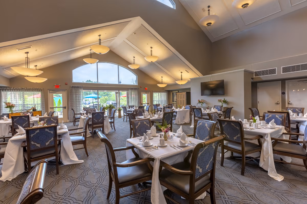 A spacious dining room with multiple tables covered in white tablecloths, each set with napkins, cups, and silverware. The room features high vaulted ceilings with hanging light fixtures, large windows letting in natural light, and a view of an outdoor patio area with umbrellas.