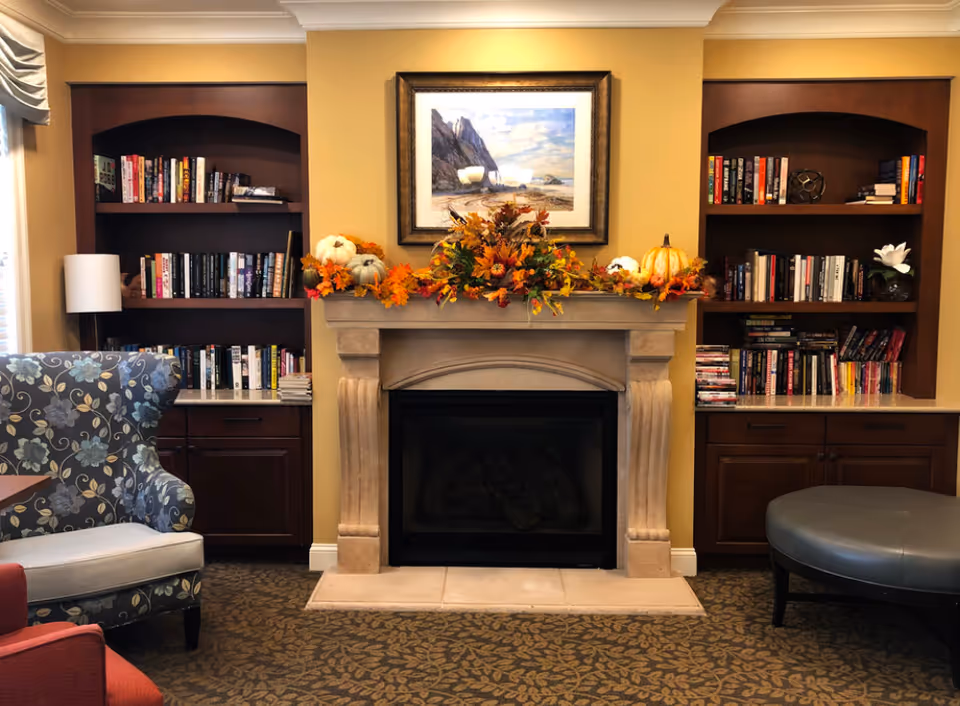 A cozy senior living room featuring a stone fireplace decorated with autumn-themed floral arrangements and pumpkins. On either side of the fireplace are built-in wooden bookshelves filled with books and decorative items. There is a floral patterned armchair on the left and a round gray ottoman on the right, all set on a carpet with a leaf pattern.