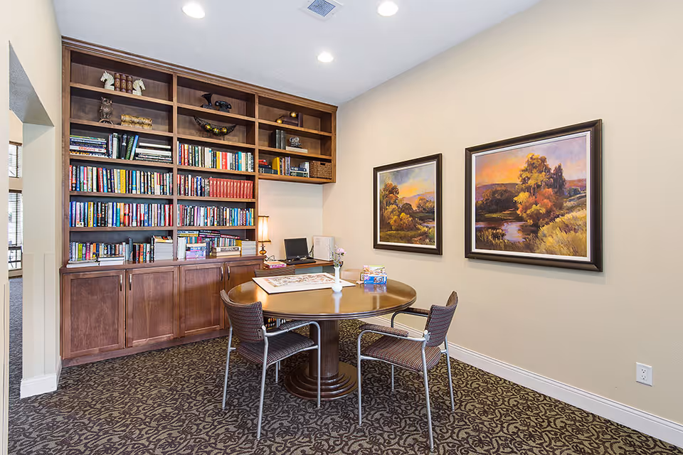 A cozy interior reading area with a round table and chairs in front of built-in bookshelves and framed landscape paintings.