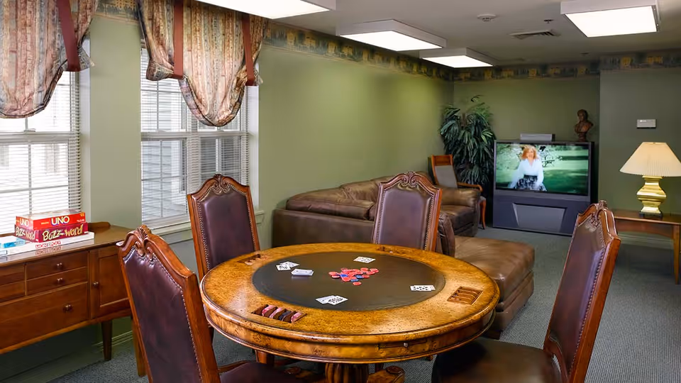 A cozy game room with a round wooden table featuring playing cards and poker chips, surrounded by four leather chairs. In the background, there is a brown leather sectional sofa, a large TV displaying a woman outdoors, a side table with a lamp, and a potted plant. The walls are painted green with patterned curtains on the windows.