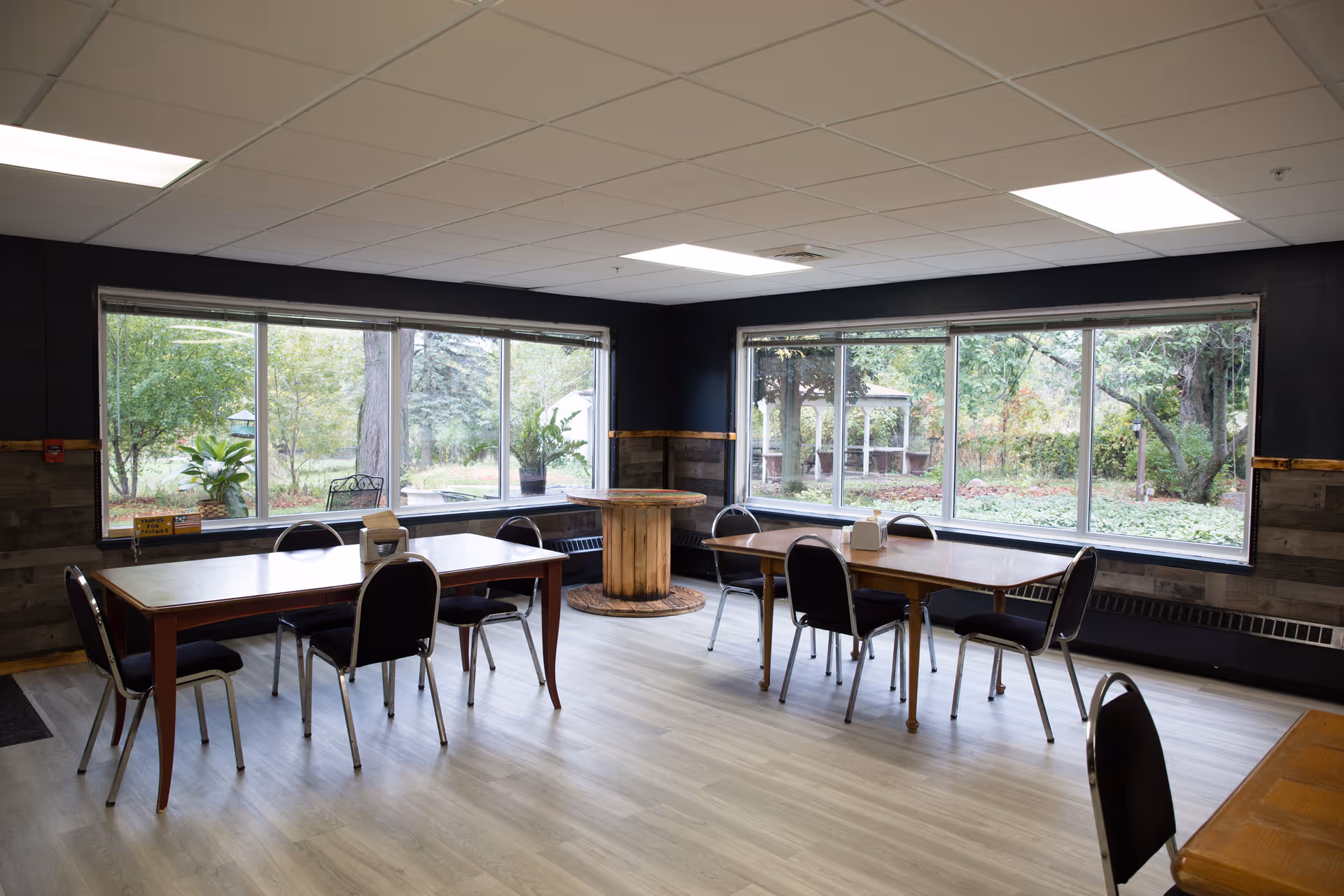 A bright communal dining room with tables and chairs and large windows looking out onto a garden and gazebo.