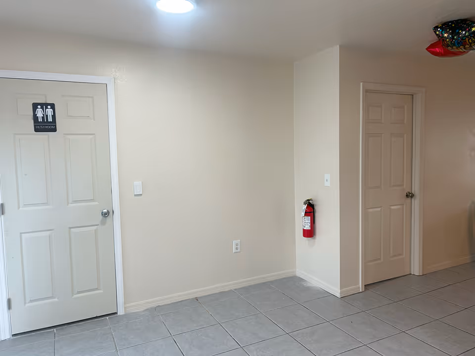 Interior room with two closed doors—one marked restroom—beige walls, tiled floor, and a wall-mounted fire extinguisher.
