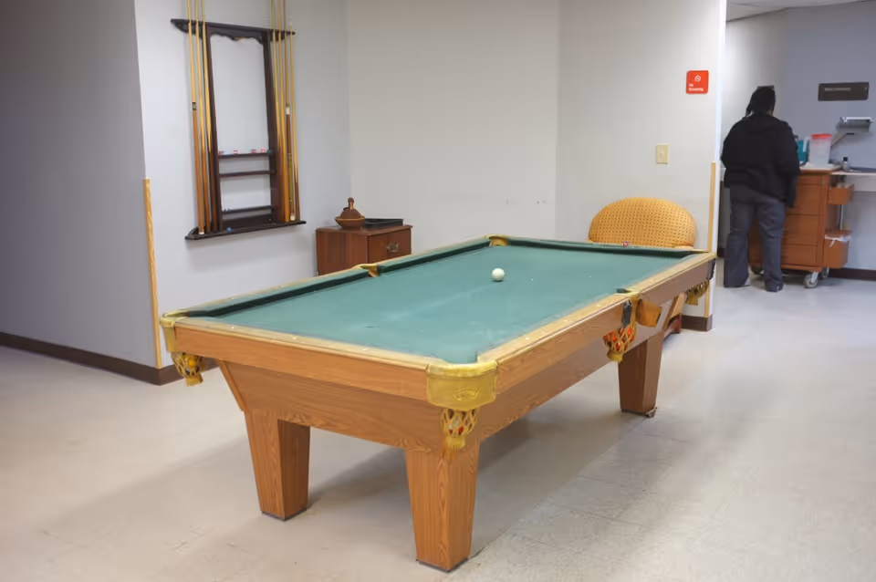 Indoor room with a pool table in the center, a rack with pool cues mounted on the wall, a small wooden cabinet, and a person standing near a counter or desk in the background. The room has plain walls and a tiled floor.