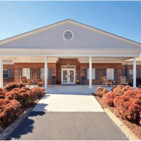 Front exterior view of a single-story brick building with white columns supporting a covered entrance. There are rocking chairs on the porch and neatly trimmed bushes lining the walkway leading to the double glass doors.