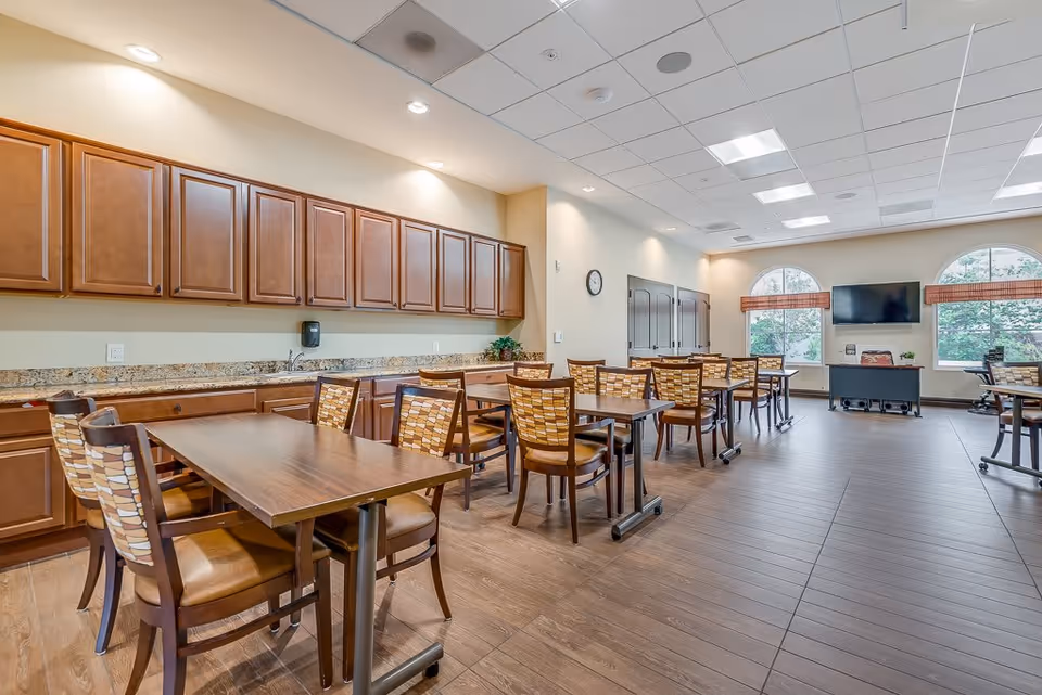 A spacious dining room with multiple wooden tables and chairs arranged neatly. The room features a long counter with wooden cabinets above and below, a granite countertop, and a sink. There are two large arched windows with blinds, a wall-mounted TV, and a clock on the wall. The floor is wood, and the ceiling has recessed lighting and ceiling tiles.