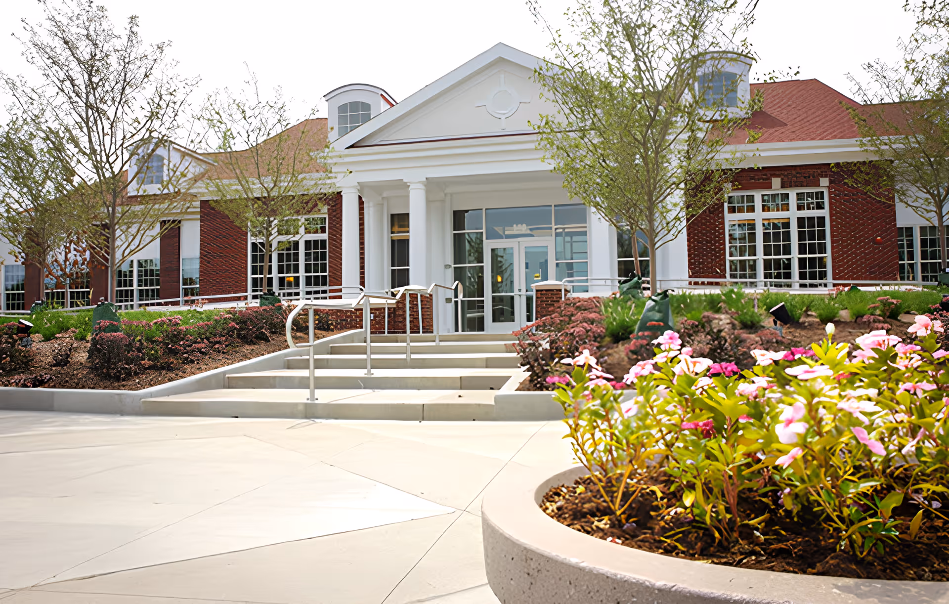 Front exterior view of Compass Park facility featuring a brick building with large windows, white columns at the entrance, steps with handrails, landscaped flower beds, and small trees.