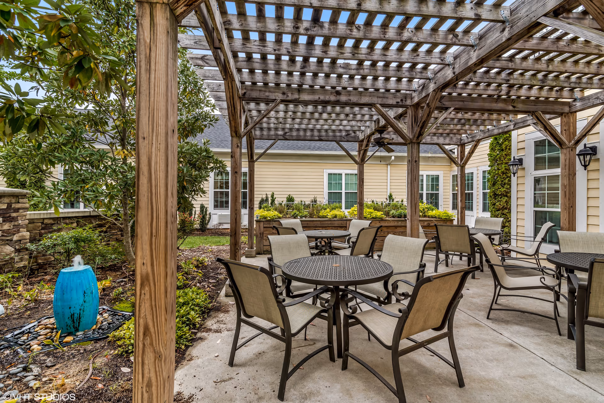Shaded outdoor courtyard with a wooden pergola, round metal tables and chairs, and a small blue fountain.