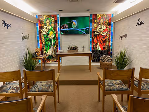 Small chapel or meditation room with wooden chairs arranged in rows facing a small altar table. Behind the altar are two stained glass windows depicting religious figures, and a flat screen TV mounted on the wall showing a bird on a branch. The walls are white brick with the words 'Hope', 'Gratitude', 'Love', and 'Forgiveness' displayed on them. There are green plants on either side of the altar and a basket with a blanket on the right side.
