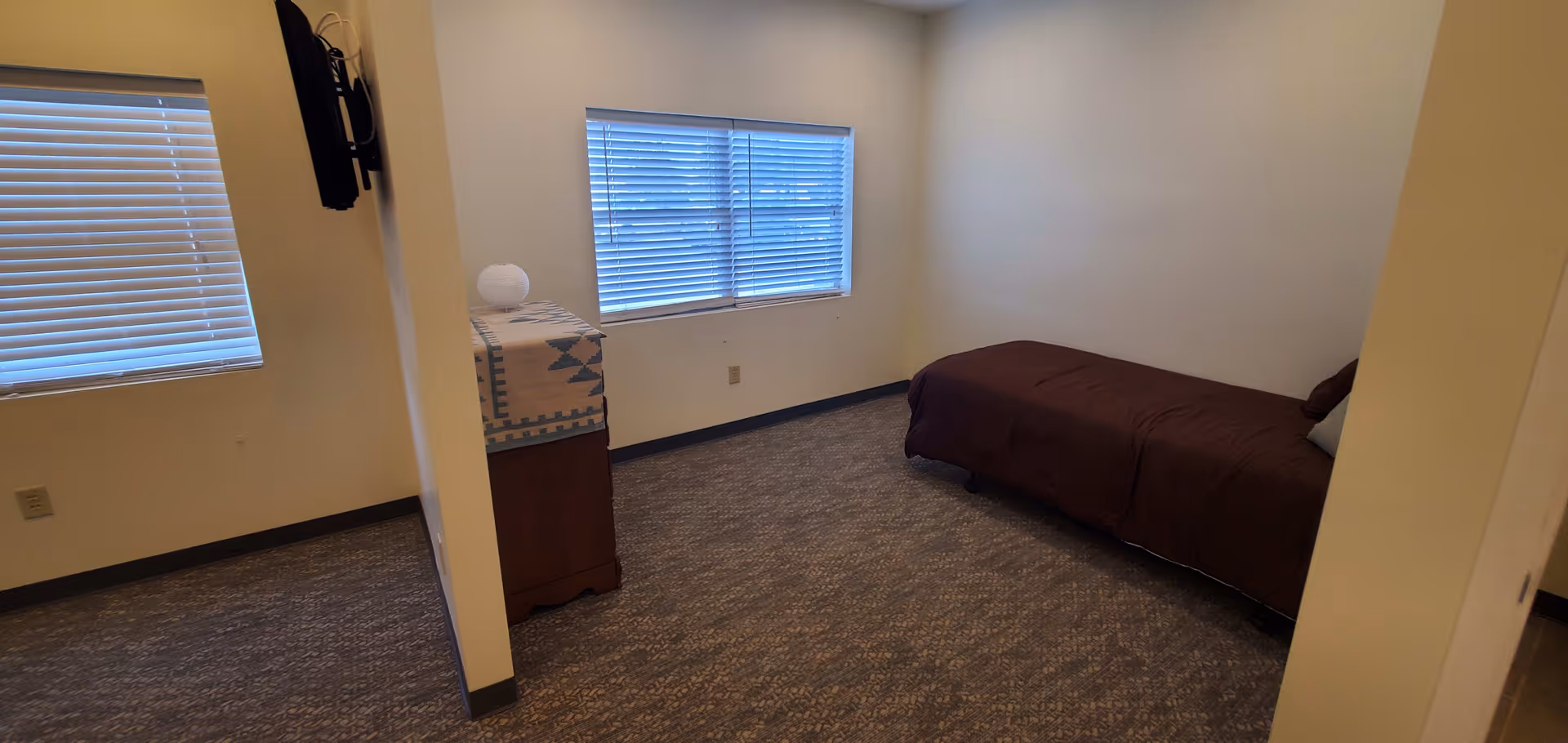 A small bedroom with a single bed covered in a dark brown blanket, a wooden dresser with a patterned cloth and a decorative item on top, two windows with closed blinds, and beige walls.