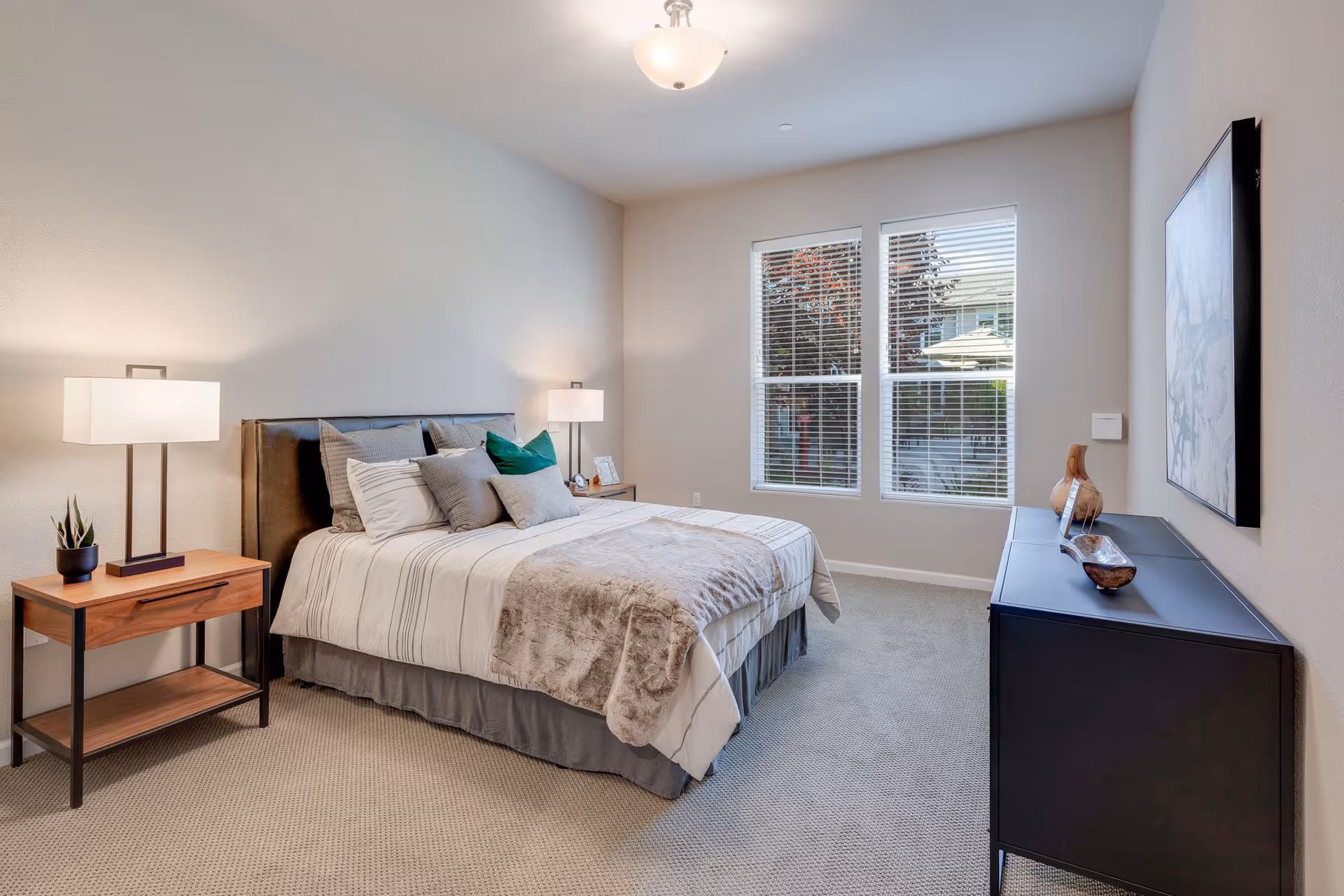 Bright, modern bedroom with a made bed, bedside tables and lamps, a dresser, and two windows.