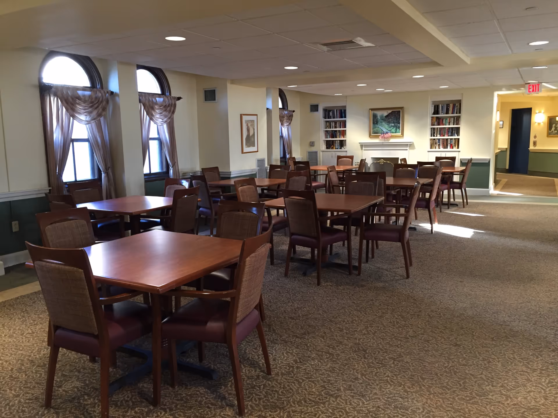 A spacious dining room with multiple wooden tables and chairs arranged neatly. The room features arched windows with sheer curtains, carpeted floor, and built-in bookshelves flanking a fireplace with a painting above it. The walls are painted in light colors with green wainscoting, and there is an exit sign visible in the background.