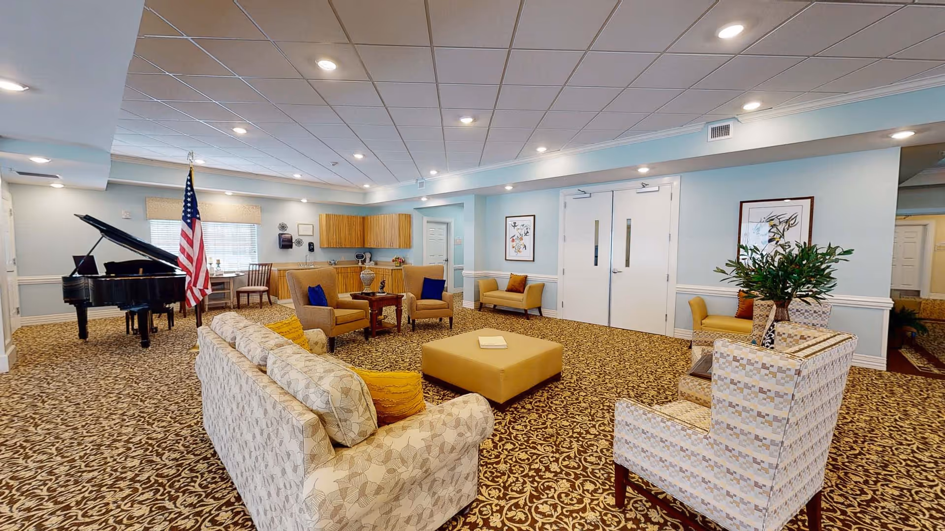 A spacious and well-lit common area with patterned carpet, multiple seating options including sofas and armchairs with cushions, a grand piano near a window with an American flag beside it, a small table with chairs, and light blue walls adorned with framed artwork and a large double door.