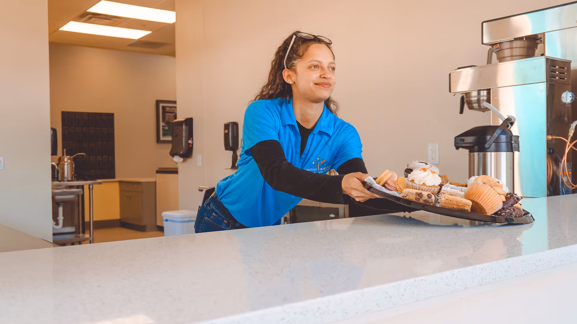 A woman in a blue shirt is placing a tray of assorted pastries and desserts on a countertop in a kitchen or serving area.