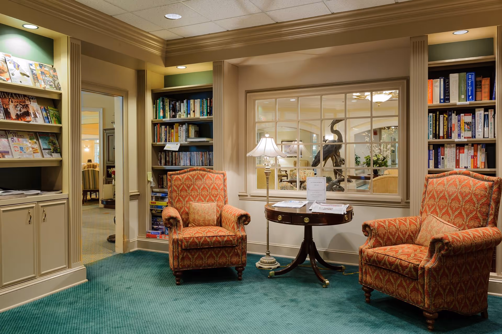 Cozy interior lounge with two patterned upholstered armchairs facing a round wooden table with a lamp, flanked by bookshelves and magazine racks.