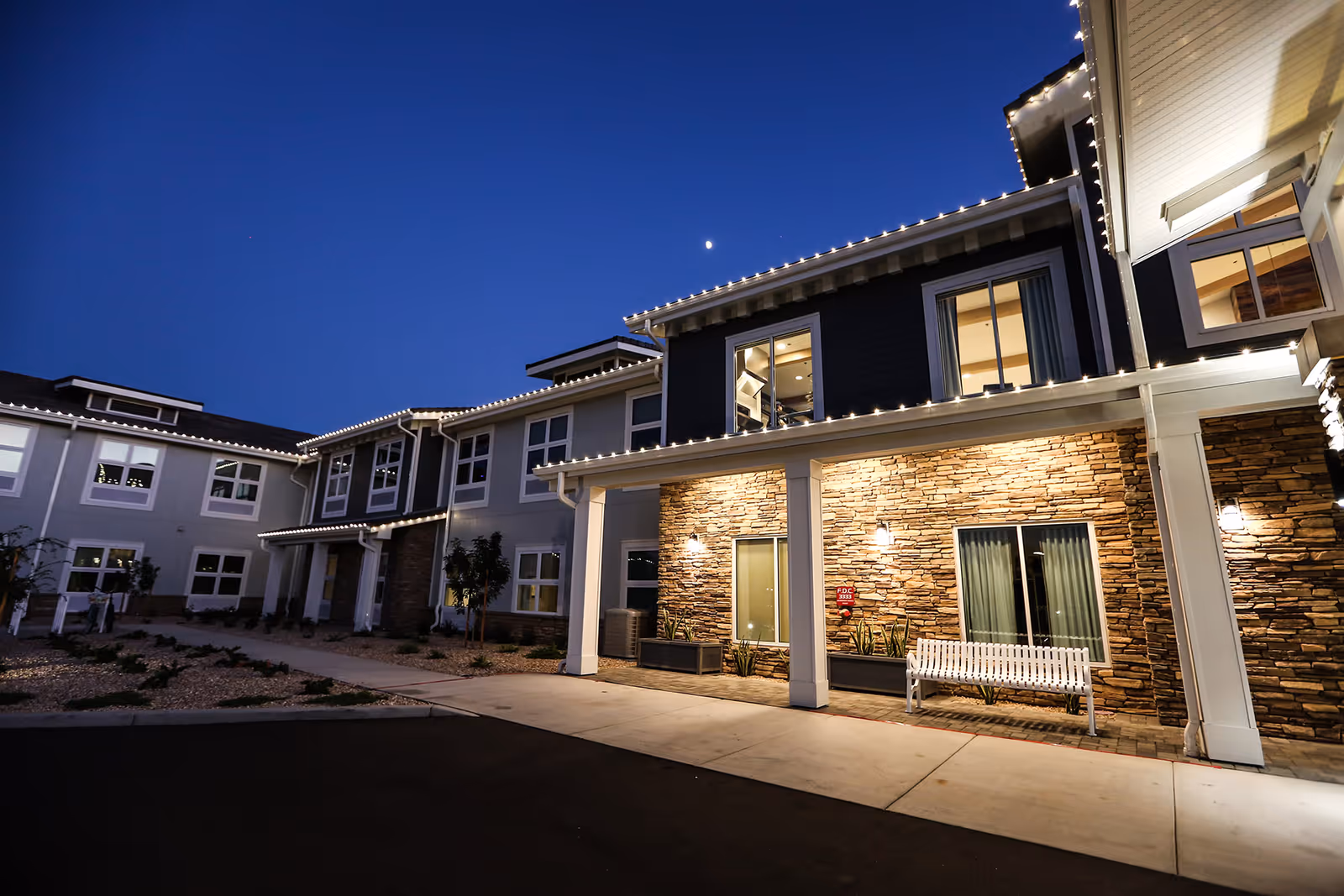 Exterior view of a senior living facility building at dusk with illuminated string lights along the roofline, stone facade, windows, a white bench, and a clear evening sky with a visible moon.