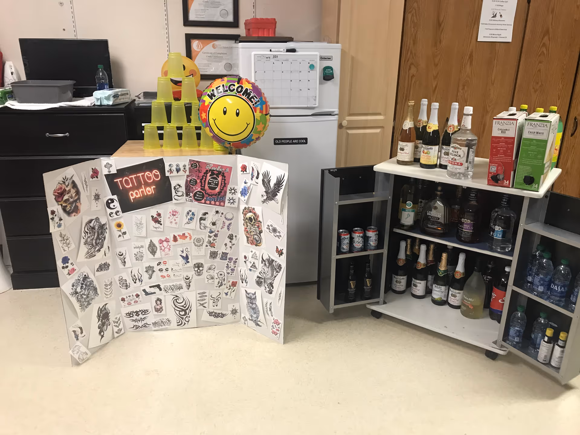 Interior scene with a folding display of temporary tattoo designs, a small fridge topped by a smiley 'Welcome' balloon and stacked yellow cups, and open cabinets stocked with bottles and drinks.