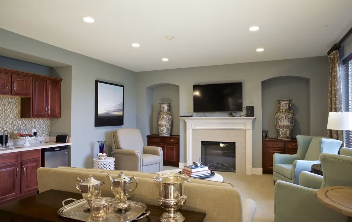 A comfortable living room with a fireplace and wall-mounted TV, upholstered chairs and sofas, decorative vases, and a small kitchenette area.