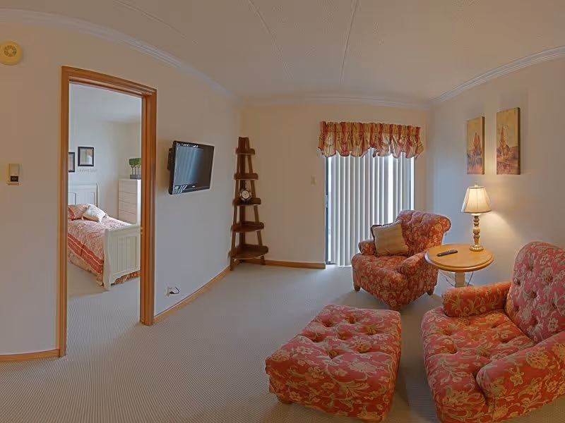 A cozy living room with floral patterned armchairs and ottoman, a small round wooden side table with a lamp, and a wall-mounted flat screen TV. There is a doorway leading to a bedroom with a white bed and dresser. Vertical blinds cover a sliding glass door, and two framed paintings hang on the wall.