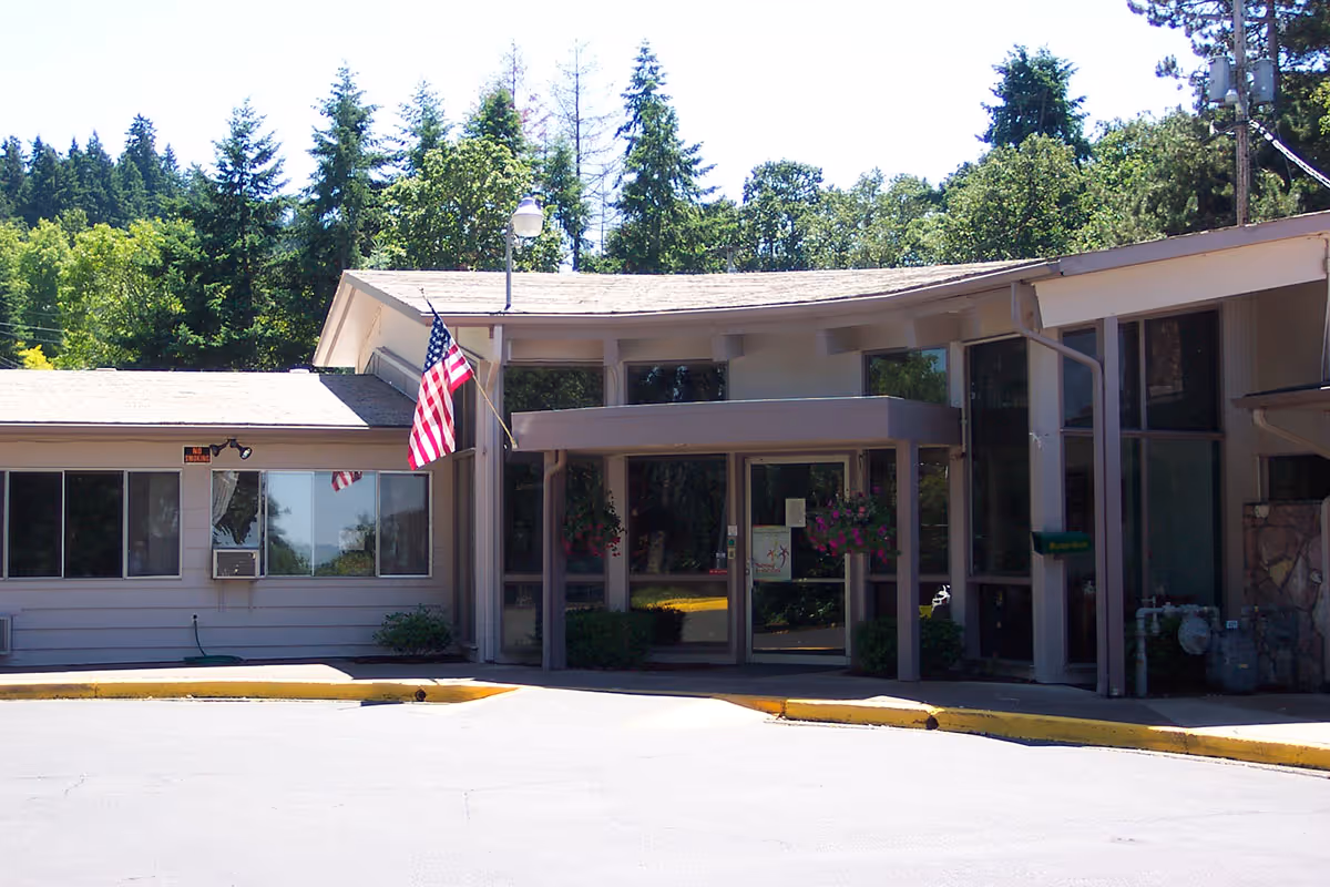 Exterior view of Hillside Heights Rehabilitation Center showing the main entrance with glass doors, an American flag mounted on a pole, hanging flower baskets, and surrounded by trees and greenery.