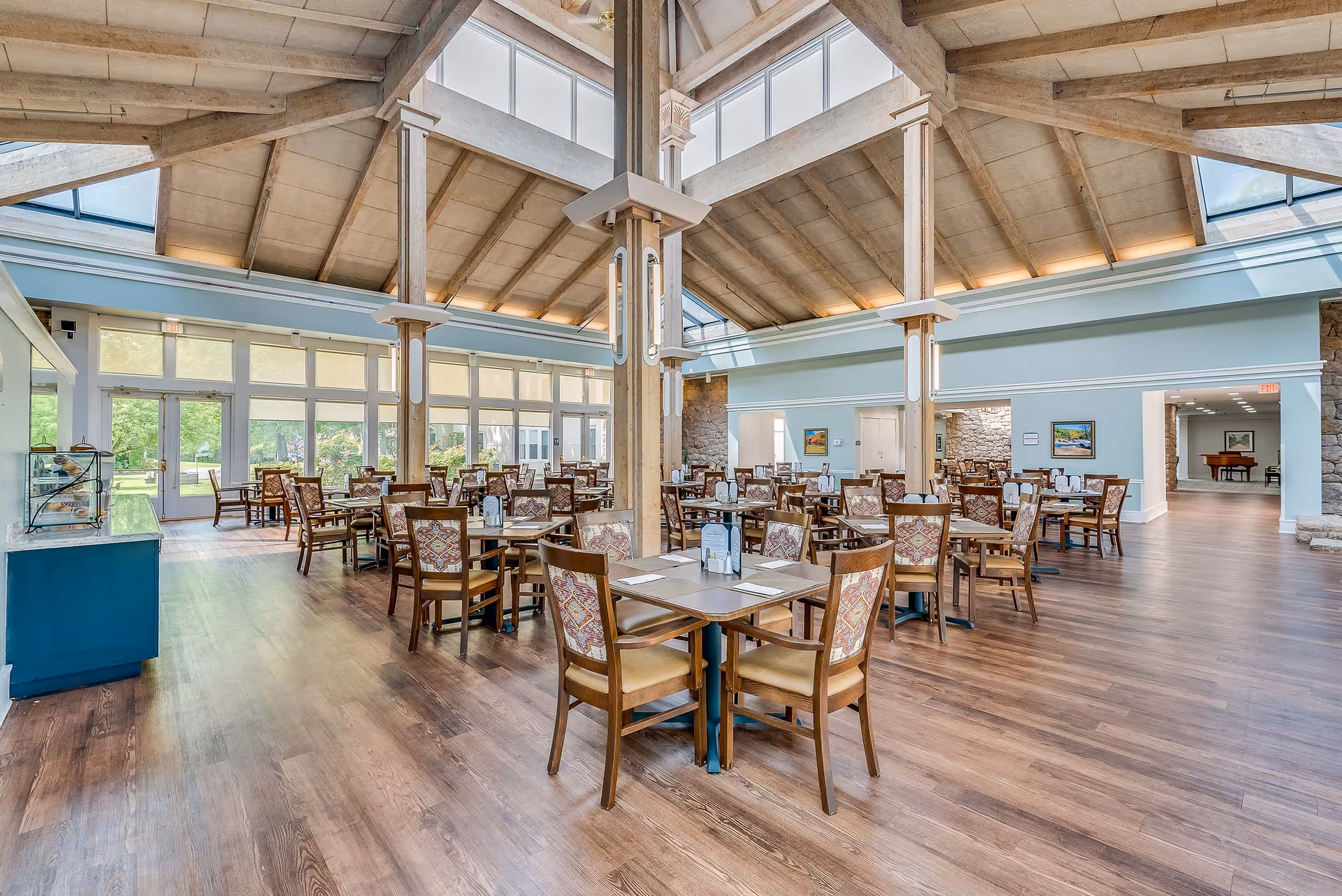 Spacious dining room with multiple wooden tables and chairs under a high beamed ceiling and large windows.