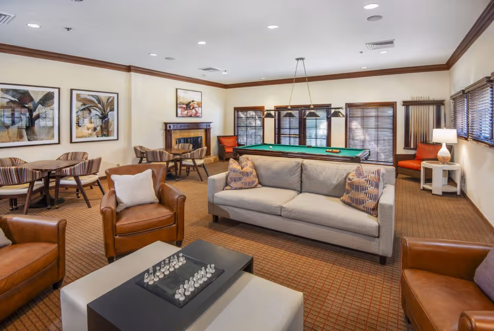 A spacious and well-lit common lounge area with a beige sofa, two brown leather armchairs, a coffee table with a chess set, and a pool table in the background. The room also features multiple small tables with chairs, framed artwork on the walls, a fireplace, and windows with wooden blinds.