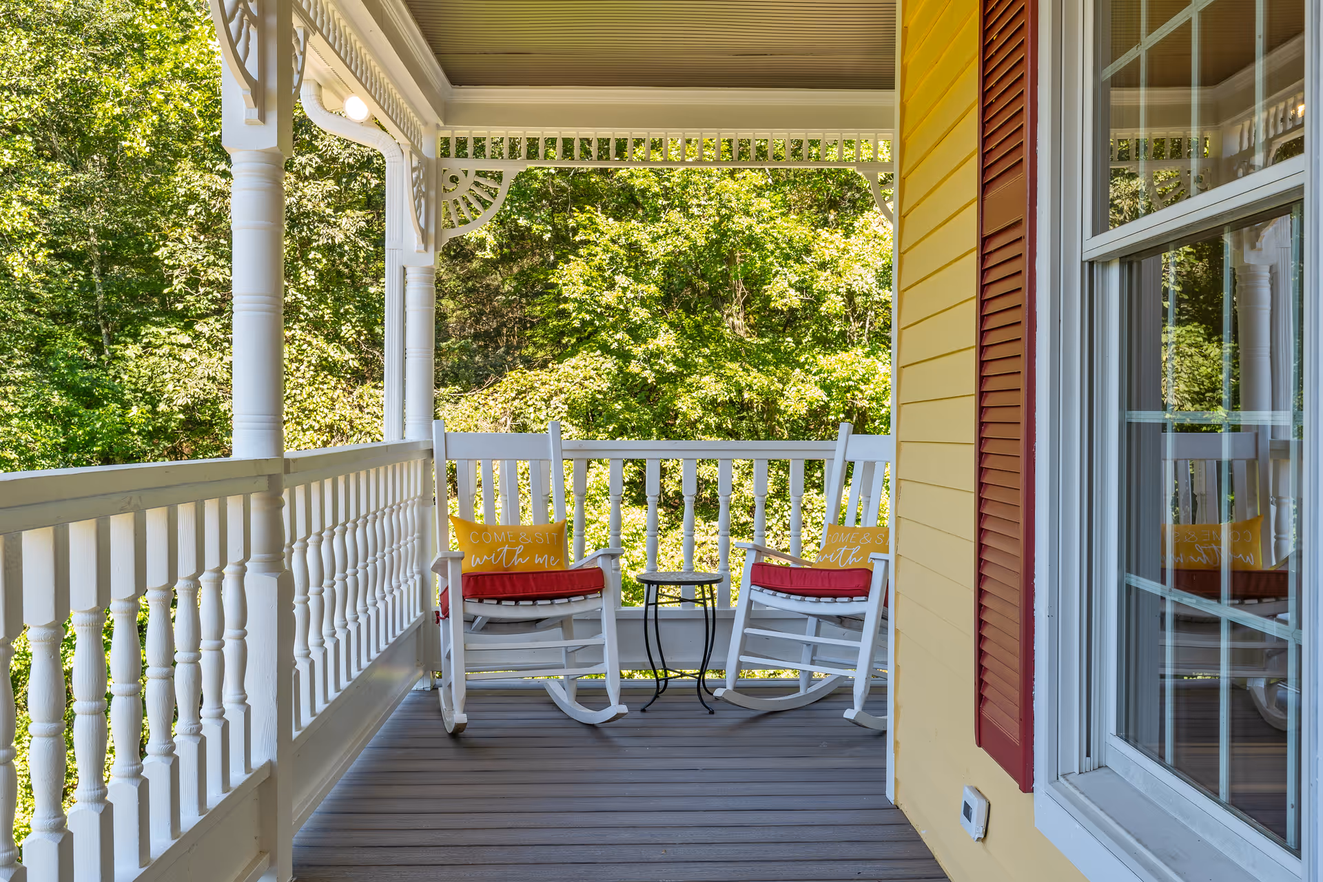A shaded wraparound porch with two white rocking chairs, a small side table, and decorative white railing overlooking green trees.