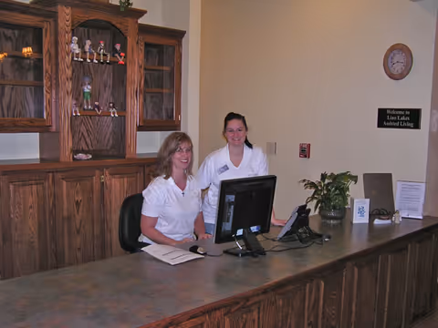 Two women in white uniforms sitting and standing behind a wooden reception desk with a computer monitor, phone, and some papers. Behind them is a wooden cabinet with decorative figurines. A sign on the wall reads 'Welcome to Lino Lakes Assisted Living.'