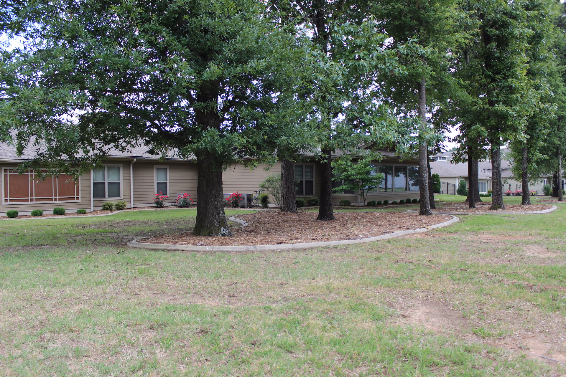 Exterior view of a single-story senior living facility building with beige siding, several windows, and a landscaped area with trees and shrubs in front. The foreground shows a grassy lawn with patches of dirt.