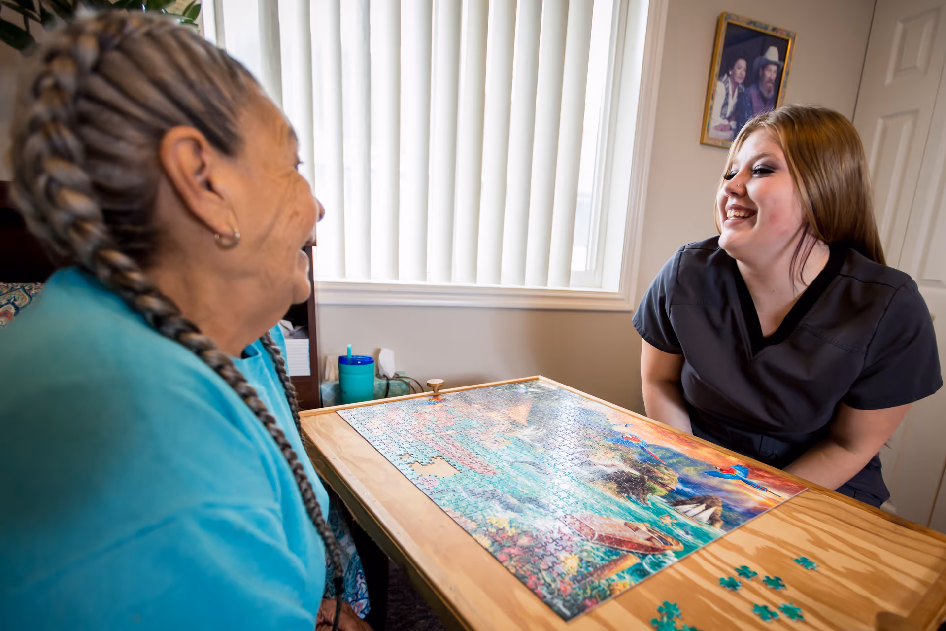 An elderly woman and a younger woman sitting at a wooden table working on a colorful jigsaw puzzle together in a well-lit room with a window and a framed picture on the wall. Both are smiling and appear to be enjoying the activity.