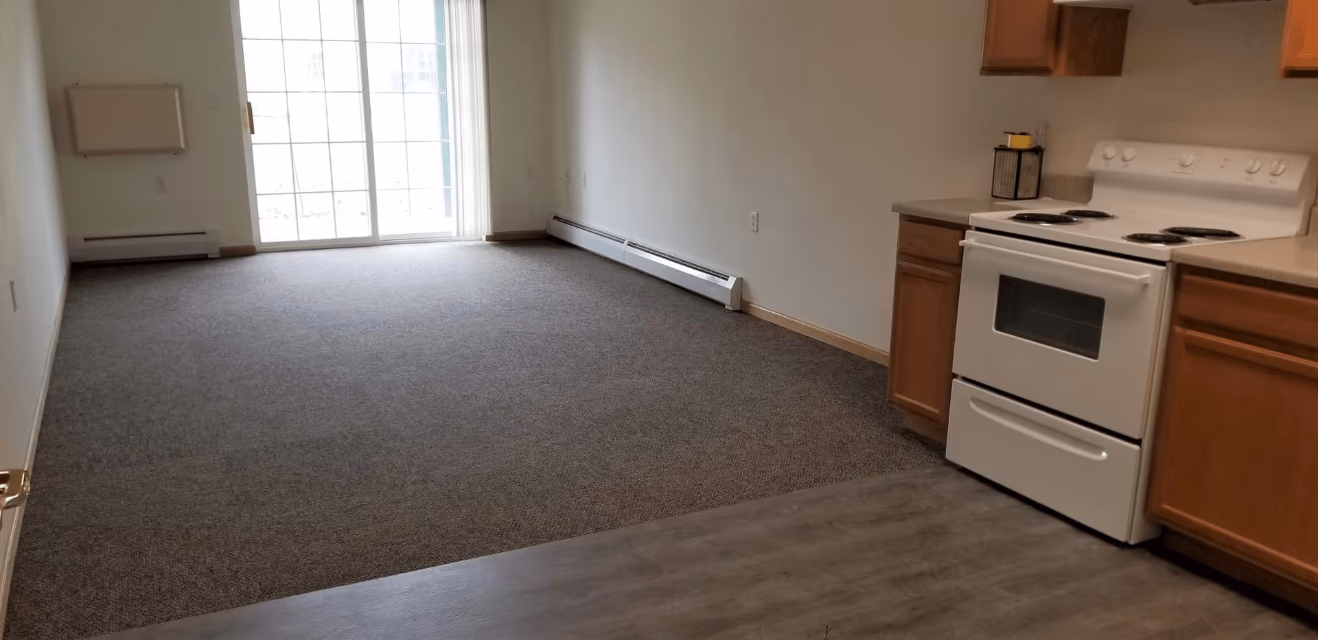 Empty room with carpeted floor and sliding glass door letting in natural light. On the right side, there is a small kitchen area with a white electric stove, wooden cabinets, and a countertop. The walls are plain and light-colored.
