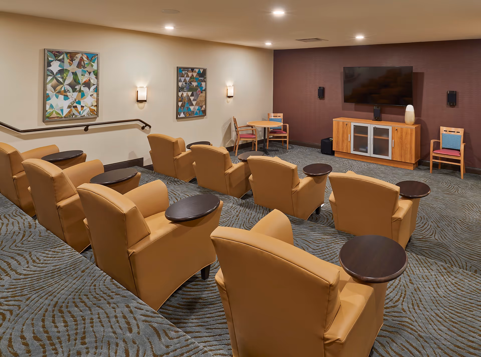 Cozy senior living media room with rows of tan recliners facing a wall-mounted TV and small side tables.