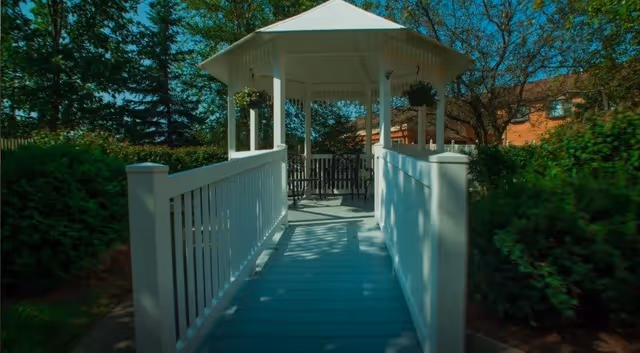 A white gazebo reached by a ramp with white railings surrounded by trees and shrubs.