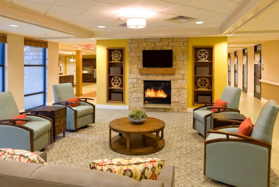 Comfortable senior living lounge with armchairs arranged around a coffee table facing a stone fireplace and wall-mounted TV.