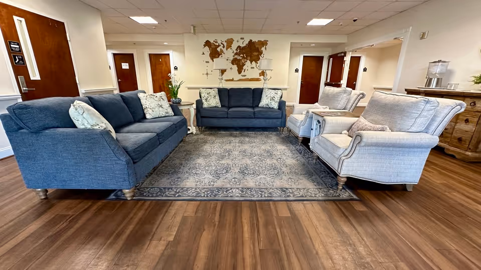 A cozy sitting area in a senior living facility with two blue sofas and two light gray armchairs arranged around a patterned area rug on a wooden floor. The walls are light-colored with wooden doors and a decorative world map wall art in the background. There is a wooden sideboard with a water dispenser on the right side.