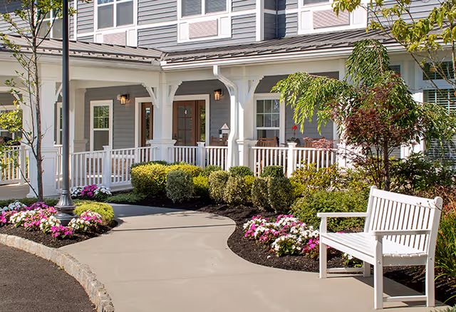 A paved walkway leading to the entrance of a senior living facility with a white railing porch, surrounded by well-maintained bushes, colorful flowers, and trees. A white bench is placed near the walkway on the right side.