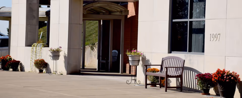 Entrance area of a building with a bench and several potted flowers arranged along the wall. The building has large windows and the number 1997 is displayed on the wall near the window.
