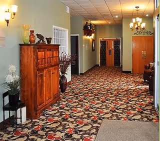 Carpeted lobby hallway with a patterned rug, wooden cabinets and decorative vases along the wall, wall sconces and chandeliers, and double doors at the far end.