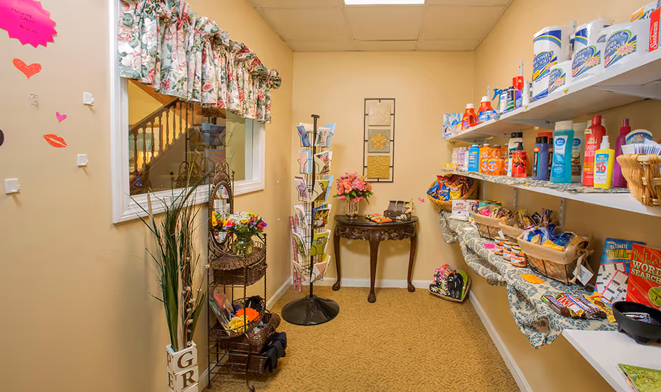 Small shop area inside a senior living facility with shelves stocked with snacks, toiletries, and household items. There is a decorative table with flowers and a rotating rack with greeting cards. The walls are beige with floral curtains on the window and some decorative wall art.